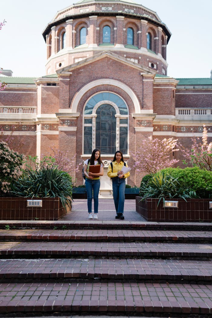Two young women walking past a historic university building on a sunny day.