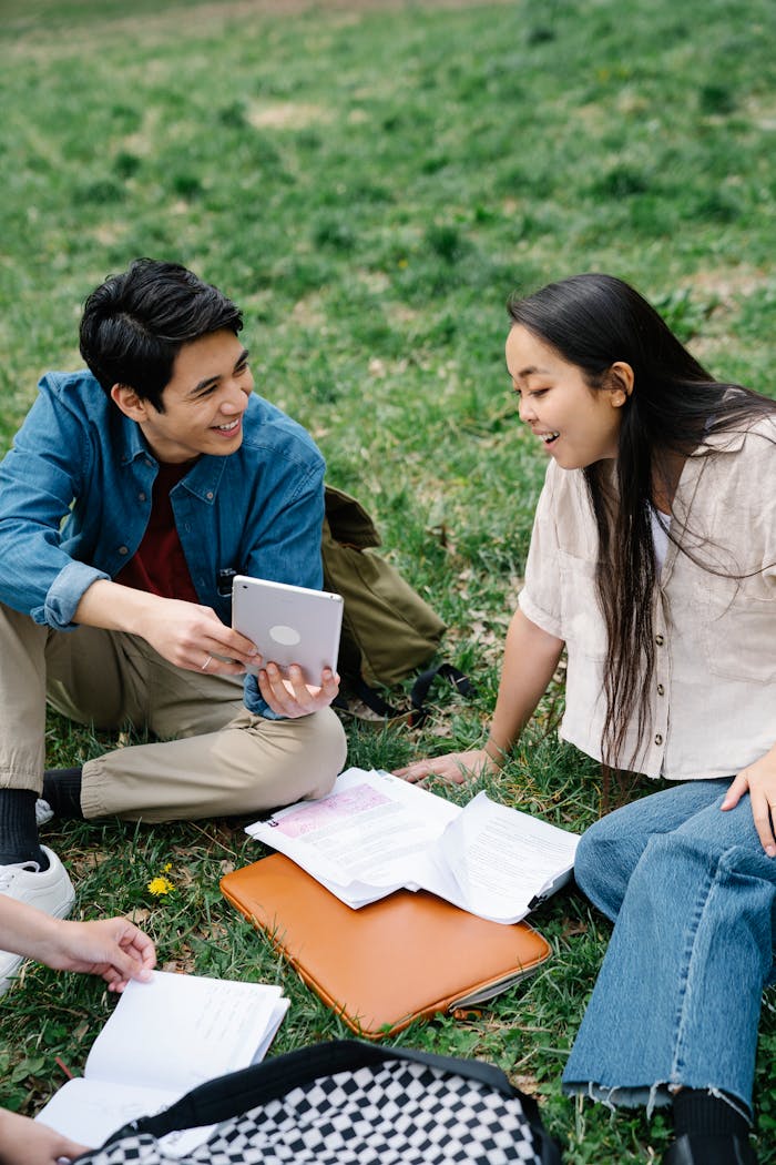 Young adults happily studying on grass, using a tablet and notes.