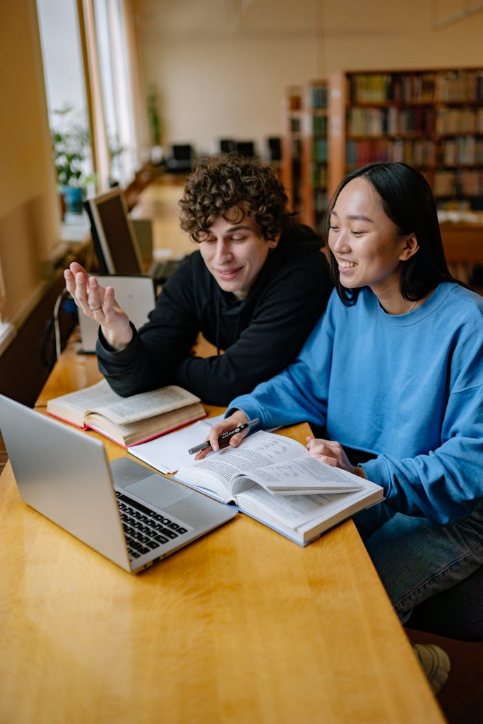 Two students study together at a library table with laptops and books.